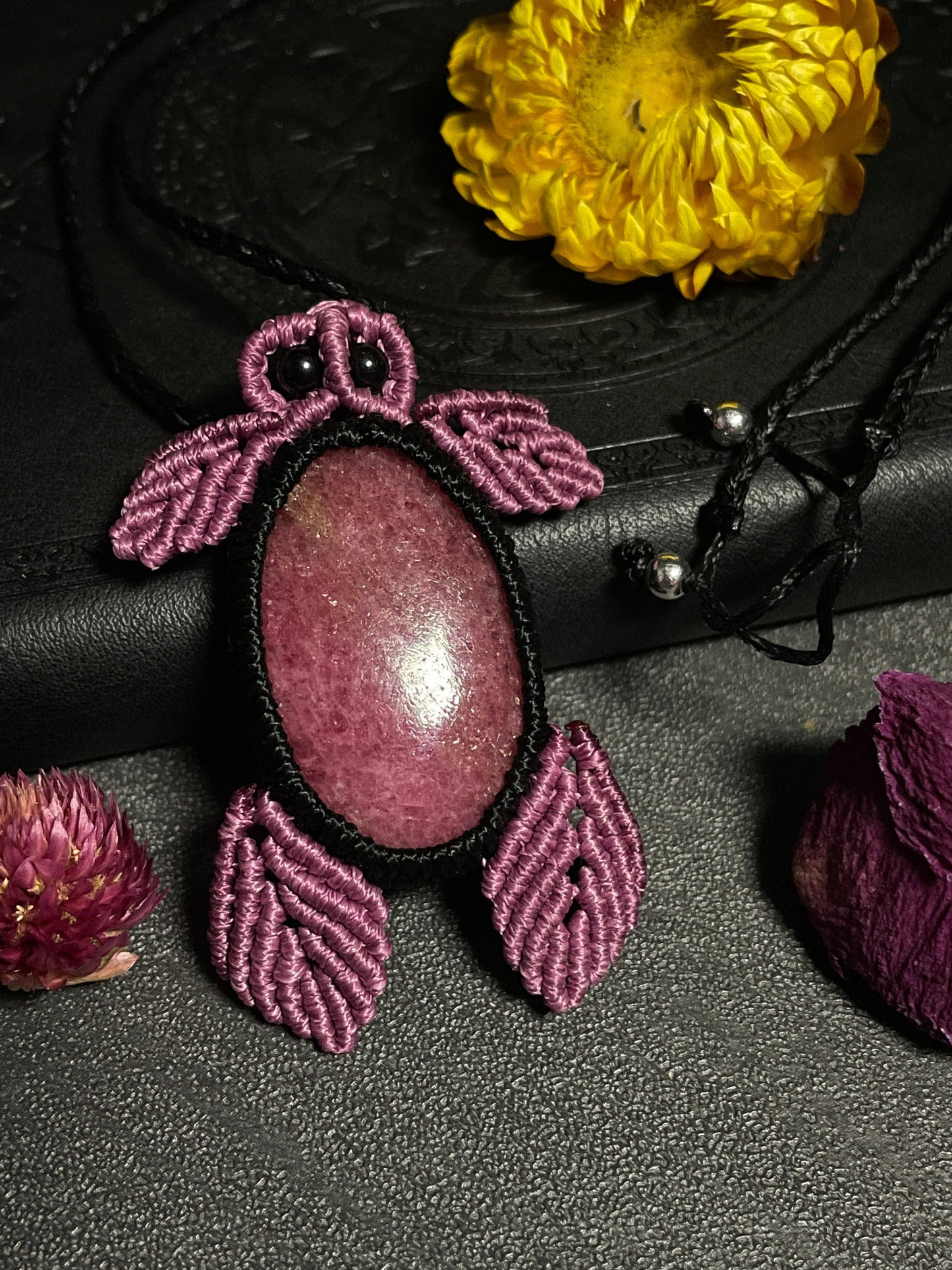 Pictured is a rhodonite cabochon wrapped in macrame thread. The rhodonite pendant is in the shape of a sea turtle. A gothic book and flowers are nearby. Rhodonite Sea Turtle Macramé Necklace (Twisted Nightshade Jewellery) Strawberry Quartz Pink Macramé Necklace (Twisted Nightshade Jewellery) - The Wandering Fox Emporium, Your Crystal Store