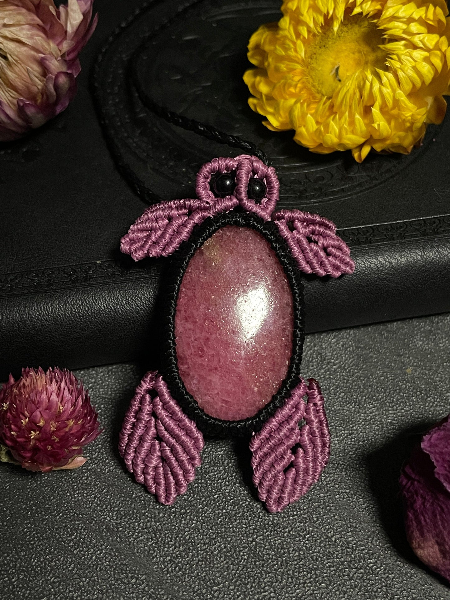 Pictured is a rhodonite cabochon wrapped in macrame thread. The rhodonite pendant is in the shape of a sea turtle. A gothic book and flowers are nearby. Rhodonite Sea Turtle Macramé Necklace (Twisted Nightshade Jewellery) Strawberry Quartz Pink Macramé Necklace (Twisted Nightshade Jewellery) - The Wandering Fox Emporium, Your Crystal Store up