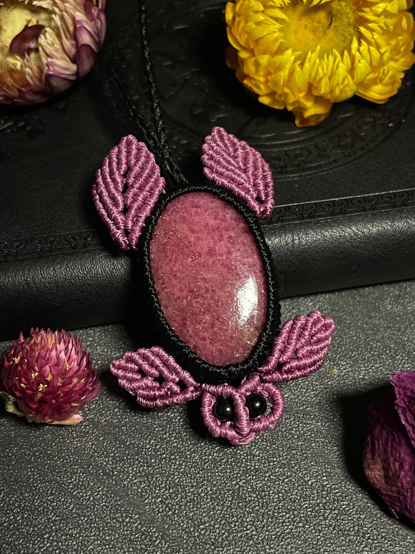 Pictured is a rhodonite cabochon wrapped in macrame thread. The rhodonite pendant is in the shape of a sea turtle. A gothic book and flowers are nearby. Rhodonite Sea Turtle Macramé Necklace (Twisted Nightshade Jewellery) Strawberry Quartz Pink Macramé Necklace (Twisted Nightshade Jewellery) - The Wandering Fox Emporium, Your Crystal Store front