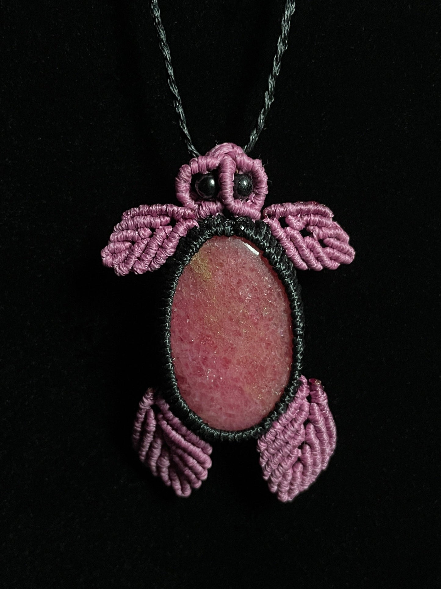 Pictured is a rhodonite cabochon wrapped in macrame thread. The rhodonite pendant is in the shape of a sea turtle. A gothic book and flowers are nearby. Rhodonite Sea Turtle Macramé Necklace (Twisted Nightshade Jewellery) Strawberry Quartz Pink Macramé Necklace (Twisted Nightshade Jewellery) - The Wandering Fox Emporium, Your Crystal Store black