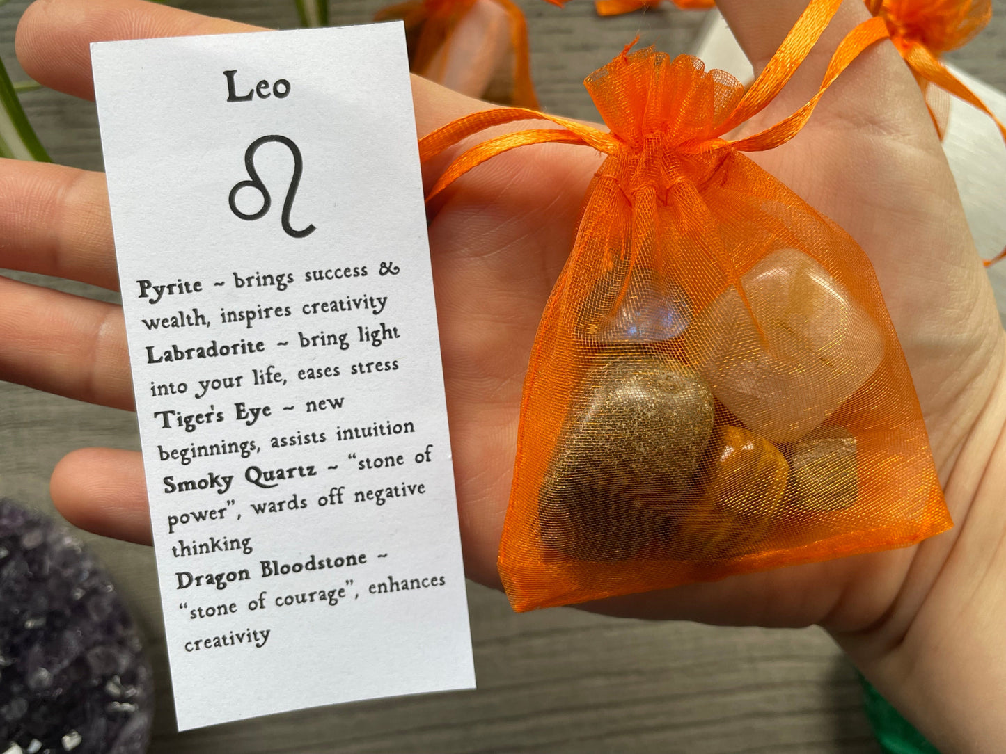 An image of a collection of tumbled stones in an organza bag. To the side is a piece of paper that describes what each tumbled stone's metaphysical properties are. LEO Zodiac Tumbled Crystal Bag close up