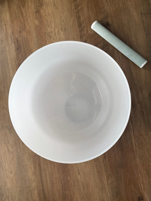 A photo of a quartz crystal singing bowl and a mallet on a wooden surface. The crystal singing bowl is made of clear quartz and has a smooth, round shape. 11" Crystal Singing Bowl (Frosted White) G top