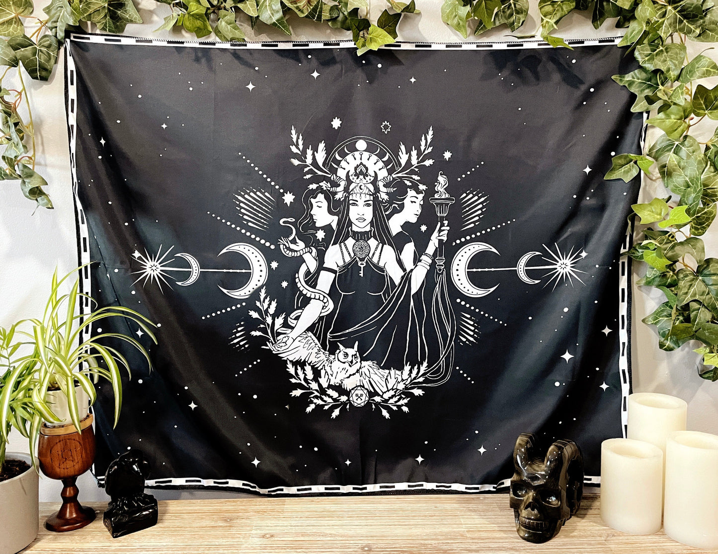 Pictured is a large wall tapestry with a black background and three women representing triple goddesses or Hecate printed in white on it. Moon Goddesses Wall Tapestry
