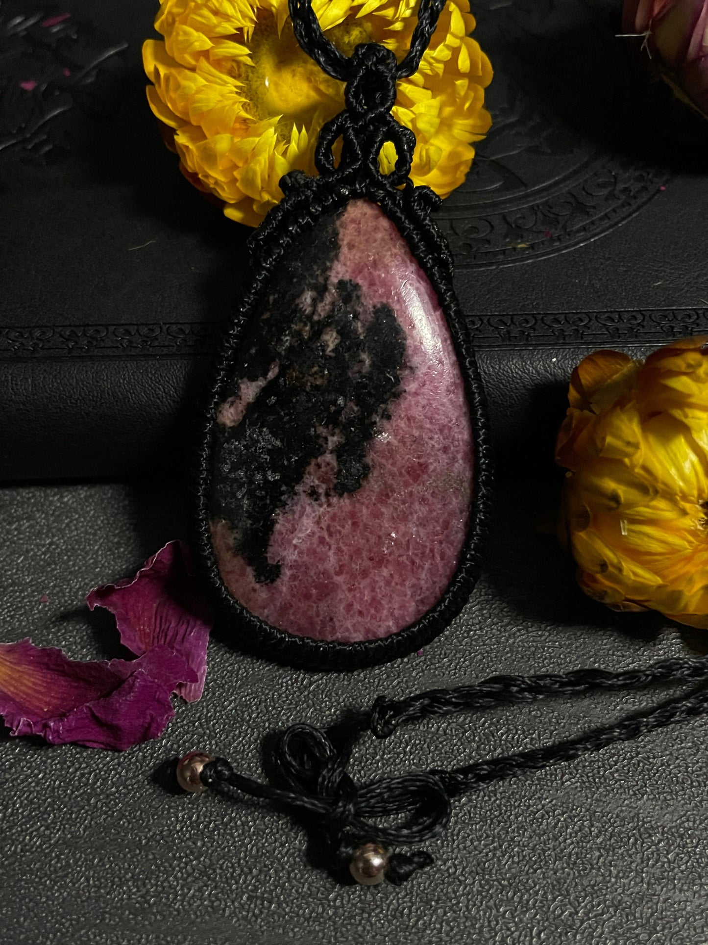 Pictured is a rhodonite cabochon wrapped in macrame thread. A gothic book and flowers are nearby. Rhodonite Macramé Necklace (Twisted Nightshade Jewellery) - The Wandering Fox Emporium, Your Crystal Store clasp