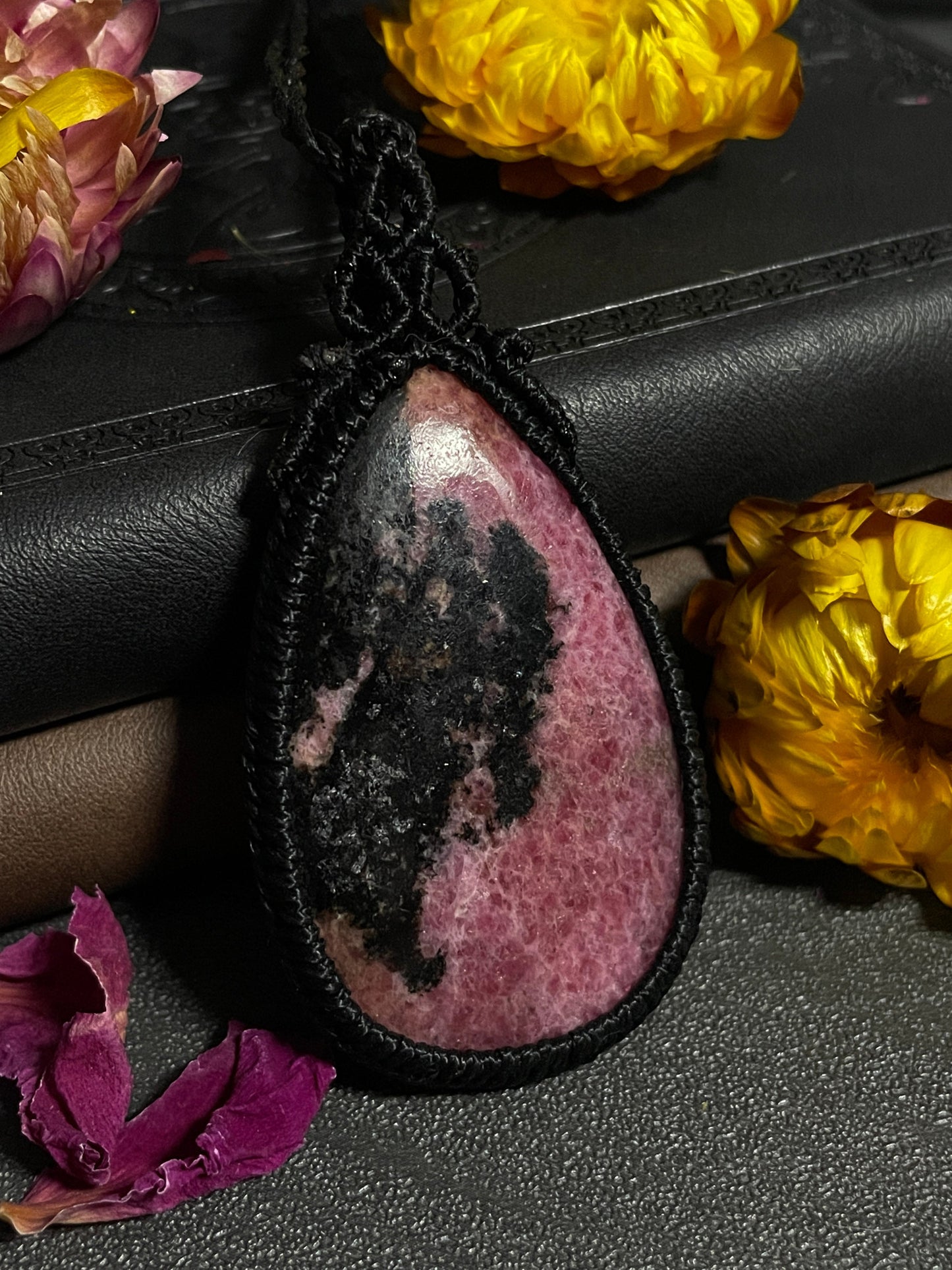 Pictured is a rhodonite cabochon wrapped in macrame thread. A gothic book and flowers are nearby. Rhodonite Macramé Necklace (Twisted Nightshade Jewellery) - The Wandering Fox Emporium, Your Crystal Store