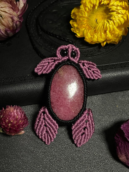 Pictured is a rhodonite cabochon wrapped in macrame thread. The rhodonite pendant is in the shape of a sea turtle. A gothic book and flowers are nearby. Rhodonite Sea Turtle Macramé Necklace (Twisted Nightshade Jewellery) Strawberry Quartz Pink Macramé Necklace (Twisted Nightshade Jewellery) - The Wandering Fox Emporium, Your Crystal Store up
