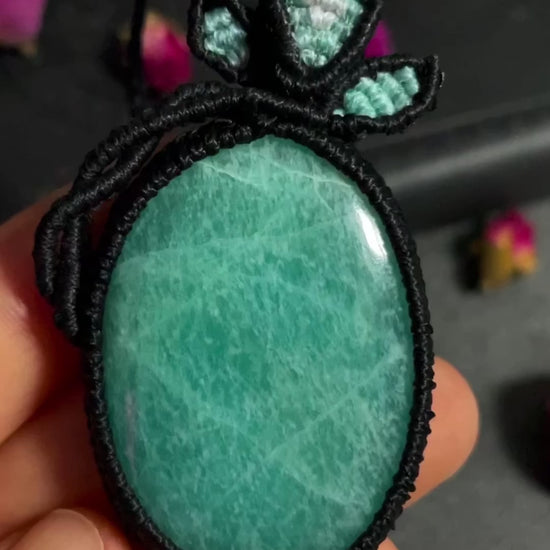 Pictured is an amazonite cabochon wrapped in macrame thread. A gothic book and flowers are nearby. Amazonite Macramé Necklace (Twisted Nightshade Jewellery) - The Wandering Fox Emporium, Your Crystal Store video
