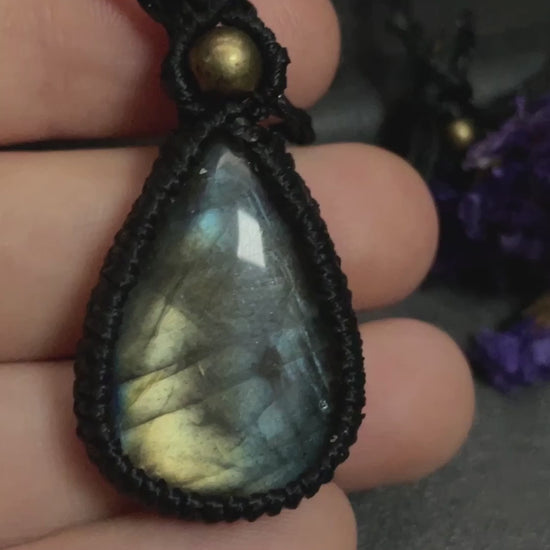Pictured is a labradorite cabochon wrapped in macrame thread. A gothic book and flowers are nearby. Blue/Gold Labradorite Teardrop Macramé Necklace (Twisted Nightshade Jewellery) - The Wandering Fox Emporium, Your Crystal Store video