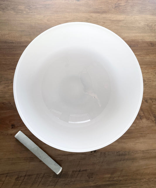  A photo of a quartz crystal singing bowl and a mallet on a wooden surface. The crystal singing bowl is made of clear quartz and has a smooth, round shape. 13" Crystal Singing Bowl (Frosted White) C4 top