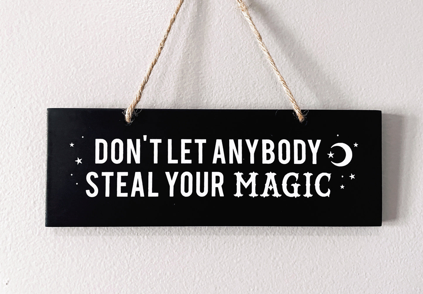 An image of a black sign with white letters that reads "Don't Let Anybody Steal Your Magic." The sign is rectangular and has a thick rope hanger attached to it, making it suitable for hanging on a wall or door.  "Don't Let Anybody Steal Your Magic" Wall Sign close up