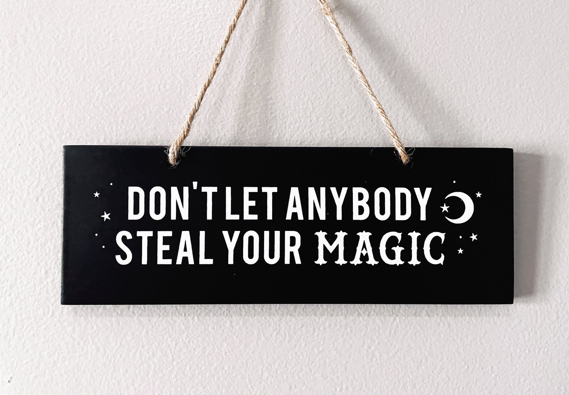 An image of a black sign with white letters that reads "Don't Let Anybody Steal Your Magic." The sign is rectangular and has a thick rope hanger attached to it, making it suitable for hanging on a wall or door.  "Don't Let Anybody Steal Your Magic" Wall Sign close up